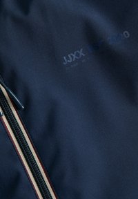 Tejido azul marino con una cremallera, con franjas rojas y blancas y texto en relieve "JXX EST. 2020" en la parte superior derecha.