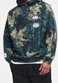 Sudadera con capucha que presenta un patrón de hojas en verde y negro, cuello con cremallera y dos logotipos: uno negro y uno rojo, en la zona del pecho.