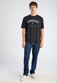 Camiseta negra con el texto blanco "LINDENBERGH 1927 COPENHAGUE", combinada con jeans de mezclilla azules y zapatillas blancas. Corte sencillo y ajuste relajado.