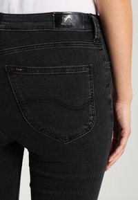 Jeans de mezclilla negra con un acabado elegante, que cuentan con un diseño de bolsillo trasero curvado y una etiqueta de cuero con la marca en la cintura.