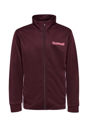 Pige smiler med armene over kors, iført en mørkerød sportjakke og leggings med pink chevronstriber, stående på hvid baggrund.