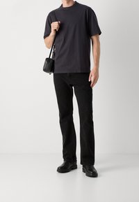 Homme portant un t-shirt noir, un jean noir et des bottes noires, tenant un petit sac bandoulière noir sur l'épaule.