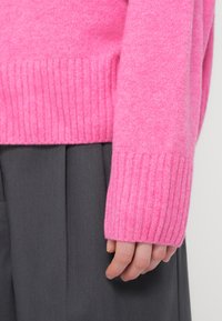 Κοντινό πλάνο ενός χεριού που καλύπτεται εν μέρει από το μακρύ μανίκι ενός ροζ πλεκτού πουλόβερ και ακουμπά δίπλα σε γκρι παντελόνια.