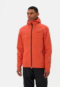 Man in een feloranje waterdichte jas met capuchon en voorrits, in combinatie met zwarte broek, staande tegen een effen witte achtergrond.