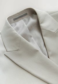 Blazer beige chiaro con un'esterno tessuto testurizzato. La fodera interna presenta un motivo a stelle grigio chiaro con una finitura liscia.