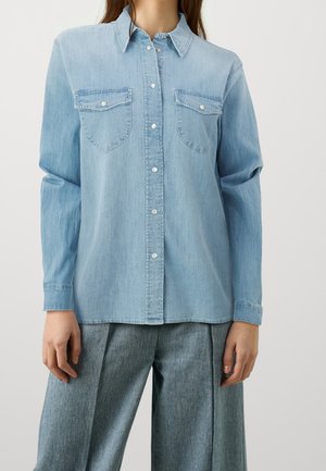 Woman wearing a light blue denim button-up shirt with two chest pockets and wide-leg blue jeans standing against a plain background.