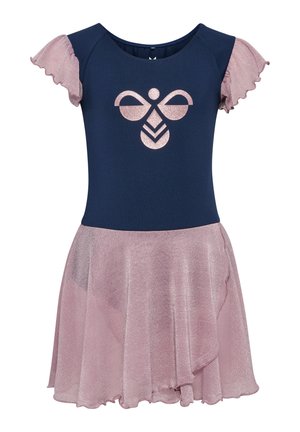 Ung pige med langt krøllet hår står med hænderne på hofterne iført en mørkeblå og pink kjole med en glitrende nederdel og sorte balletflade sko.