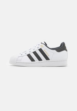 Baskets blanches Adidas Superstar avec des bandes noires, œillet doré, bout coquille et col de talon rembourré gris sur fond blanc.