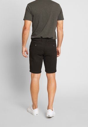 Man draagt een donkergrijs T-shirt, zwarte korte broek met knopen en witte sneakers, staand met zijn rug naar de camera tegen een effen lichte achtergrond.