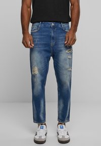 Vaqueros para hombre con efecto desgastado en mezclilla azul medio, corte relajado con desgaste, detalle de desgastes en las rodillas, combinados con zapatillas blancas y grises.