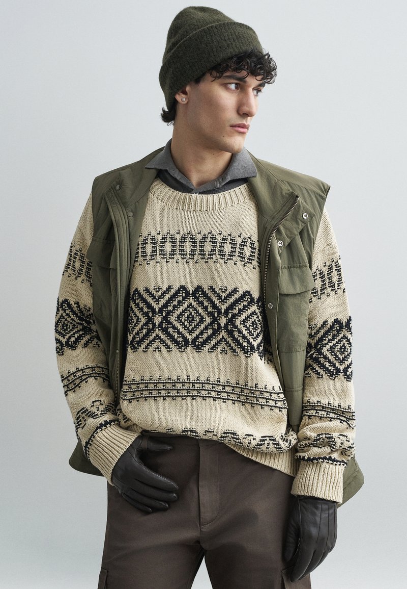 Beige kötött pulóver fekete geometrikus mintákkal, olajzöld mellény, szürke galléros ing, barna nadrág és fekete bőr kesztyű.