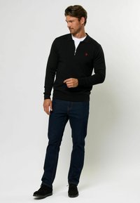 Črn zip up pulover s ovratnikom, ki ima majhen rdeč logotip na levem prsnem delu. V kombinaciji z temno modrimi kavbojkami in črnimi čevlji.
