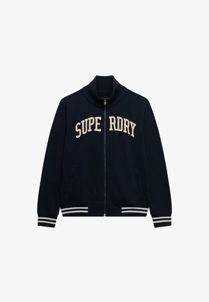 Marineblå jakke med lynlås og høj krave, beige "SUPERDRY"-tekst på brystet og hvide stribede manchetter og kant. To forlommer.