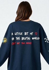 Felpa della marina con vestibilità over-size. Presenta varie grafiche e testi colorati sulla parte posteriore, incluso un design con sole e cuore, in caratteri audaci.