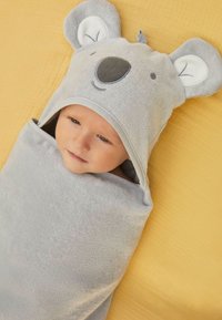 Toalla para bebé de temática de koala gris con diseño de orejas y cara, textura de toalla suave, envolviendo a un bebé, colocada sobre una superficie amarilla.