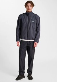 Mörkgrå fleecejacka med hög krage och dragkedja framtill, tillsammans med matchande mörka byxor. Båda har diskreta logotypdetaljer.