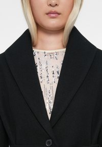 Blazer de lana negra con solapas anchas, que presenta un cierre de botón. Debajo, una blusa con lentejuelas y una superposición de tela transparente.