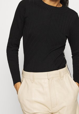 Personne portant un pull noir côtelé à manches longues rentré dans un pantalon beige taille haute, les mains dans les poches.