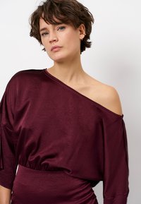 Bordeaux off-shoulder top van gestructureerde stof, ruime pasvorm en lange mouwen. Zachte, gedrapeerde vorm benadrukt de decolleté.