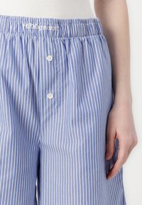 Zbliżenie na pasiastą, niebiesko-białą piżamę w postaci szortów z guzikami i elastycznym pasem, na dolnej części tułowia i ramieniu osoby.