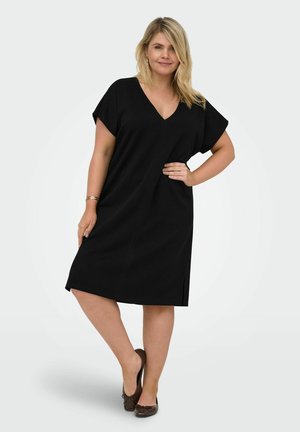 Kobieta o blond włosach, ubrana w czarną sukienkę za kolano z dekoltem w szpic i brązowe baleriny, stojąca z jedną ręką na biodrze na jednolitym tle.