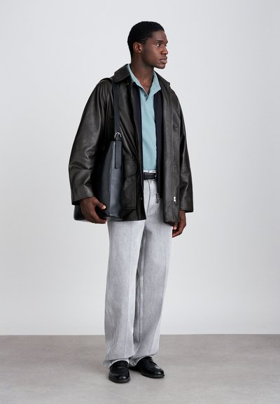 Melna ādas virsjaka, tirkīza krāsas polo krekls, pelēkas platās bikses un melnas kurpes. Tur rokās tumšu somu ar siksnu. Gludas tekstūras, moderns dizains.