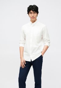 Balti medvilniniai marškiniai su ilgomis rankovėmis, turintys krūtinės kišenę; derinami su tamsiai mėlynomis kelnėmis, paprastas ir švarus dizainas.