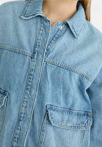 Lichtblauwe denim shirt met een klassieke kraag, knoopsluiting en twee voorzakken. Kenmerkt zich door een relaxte pasvorm en zichtbare stikseldetails.