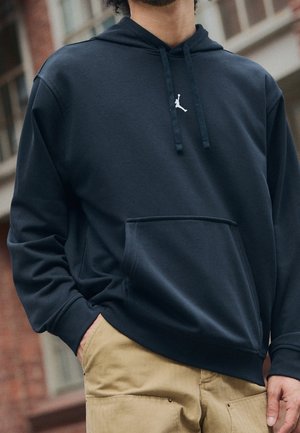 Person trägt einen schwarzen Hoodie mit kleinem weißem Jumpman-Logo und beige Hose, steht draußen mit einer Hand in der Tasche.