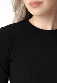 Juodas ilgomis rankovėmis viršus su apvaliu iškirpimu. Audinio tekstūra yra lygi su vertikaliais dryžiais. Minimalistinis dizainas be pastebimų akcentų.