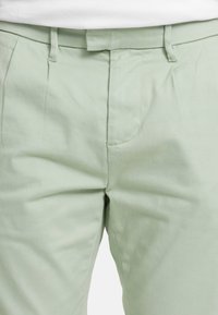 Pantalon vert clair avec passants pour ceinture et braguette zippée à l'avant, montré de la taille jusqu'à la mi-cuisse.