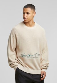 Beige ribbet sweater i løs pasform med rund hals og broderet tekst i grøn foran. Blød tekstur med lange ærmer.