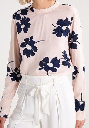 Kobieta ubrana w jasnoróżową bluzkę z długim rękawem, ozdobioną dużymi granatowymi kwiatowymi wzorami, w połączeniu z białymi, wysokim stanem spodniami przewiązanymi z przodu kokardą.