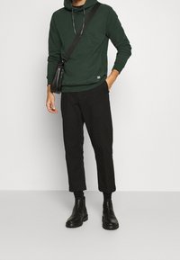 Hombre vestido con una sudadera oscura verde, pantalones negros cortos, botas de cuero negras hasta el tobillo, y una bolsa negra cruzada, frente a un fondo liso.