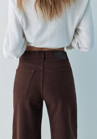 Személy magas derekú sötétbarna nadrágot és rövidített fehér hosszú ujjú felsőt visel, hátulnézetben egyszínű világos háttér előtt.