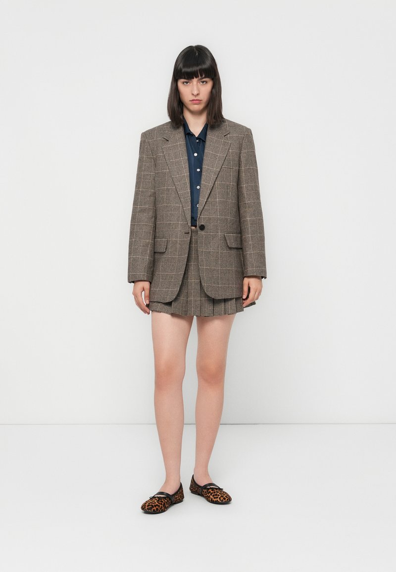Blazer oversize a scacchi marrone, camicia blu con bottoni, pantaloni corti beige a pieghe e scarpe flat con stampa leopardo. Texture morbide, design strutturato.