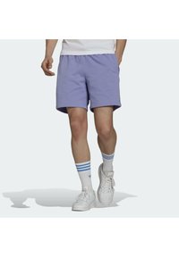 Osoba v světlých fialových šortkách, bílých teniskách a bílých ponožkách s modrými pruhy, kráčející proti jednoduchému světlému pozadí.