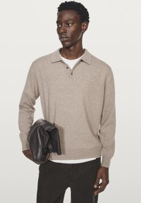 Beige stickad tröja med polokrage, tre knappar och lång ärm. Modellen håller en mörkbrun läderjacka. Vit bakgrund.