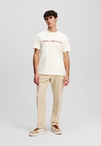 Crème katoenen t-shirt met "KARL LAGERFELD" in bruin gedrukt; gecombineerd met beige broek en wit met bruine sneakers.