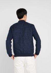 Námořnická bombardovací bunda s texturovaným povrchem, žebrovými manžetami a lemem a jednoduchým límcem. Vyznačuje se čistým, minimalistickým designem.