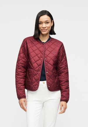 Frau trägt eine kastanienbraune gesteppte Jacke mit Knöpfen, navy Top und weiße Hose, steht vor einem einfarbigen hellgrauen Hintergrund.