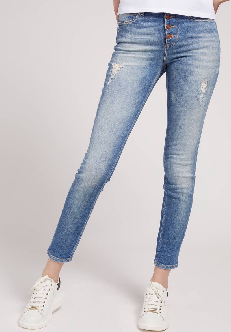 Jeans skinny azules claros con detalles desgastados, cierre de botones, cinco bolsillos y costuras en contraste; combinados con zapatillas blancas.