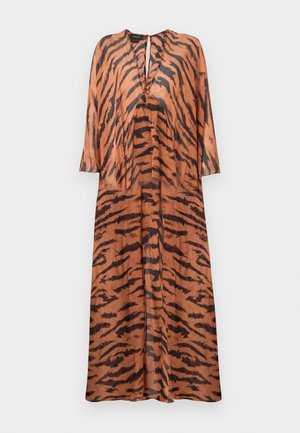 Kobieta ubrana w lejącą się sukienkę w brązowo-czarne paski tygrysa z głębokim dekoltem w kształcie litery V oraz czarne sandały na wysokim obcasie, stojąca na jednolitym tle.