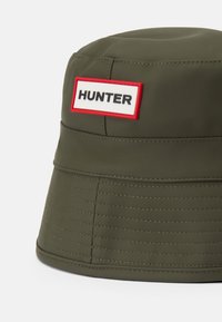 Zelenohnedý vodoodolný klobúk s okrajom v tvare vedra s horizontálnym prešívaním, s obdĺžnikovým bielym štítkom s nápisom "HUNTER" v čiernej farbe a červenými akcentmi.