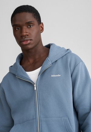 Sweatshirt azul com fecho éclair, feito de tecido suave, apresentando um bolso frontal e um logótipo bordado. Inclui capuz com cordão e punhos canelados.