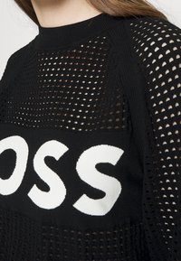 Svart stickad tröja med en lös nätdesign, med en framträdande vit "BOSS"-logga tvärs över mitten och en ribbad rund halsringning.