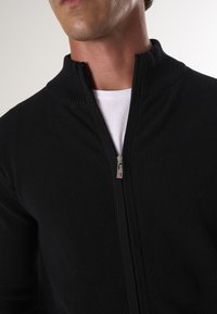 Zwarte ritssweater met een geribbelde textuur en hoge kraag. Voorzien van een metalen rits en een contrasterend wit T-shirt eronder.