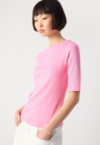 Jonge vrouw met kort donker haar die een roze, aansluitend T-shirt met ellebooglengte mouwen en witte broek draagt, met haar handen in de zakken, tegen een effen achtergrond.