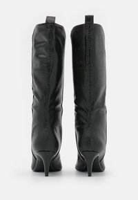 Botas negras hasta la rodilla con un acabado texturizado, punta afilada y tacón de aguja delgado. Cuenta con una pestaña en la parte posterior para facilitar su uso.