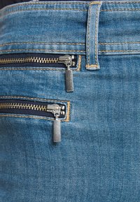 Modré denimové džíny s dvěma ozdobnými zipovými kapsami se stříbrným kováním. Látka má texturovaný povrch s zlatými švy.